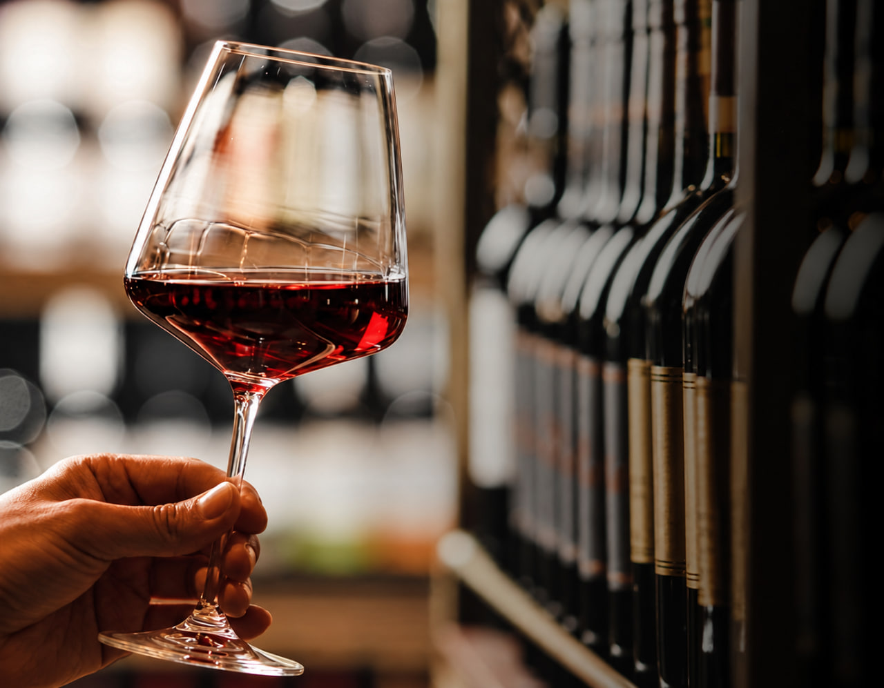 Verre de vin rouge tenu devant des bouteilles en cave